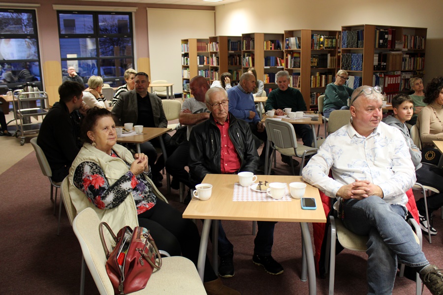 Osoby z publiczności siedzą przy stolikach i w skupieniu słuchają prelekcji autora.