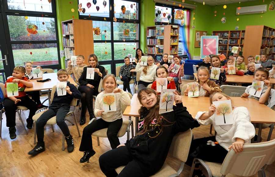 Dzieci siedzą przy stolikach a w rękach trzymają wykonane prace którymi są obrazki drzew w jesiennych kolorach.