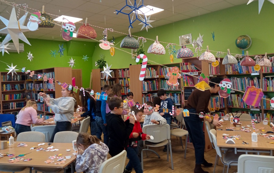 Dzieci stoją i zakładają ozdoby na sznurek lub siedzą przy stołach i pracują