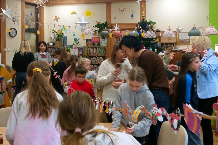 Dzieci, bibliotekarki i nauczyciele zgromadzeni w oddziale dla dzieci i młodzieży tworzą kolorowy łańcuch z ozdób świątecznych