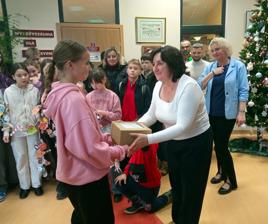 Dyrektorka biblioteki wręcza prezent na ręce jednej z uczennic odwiedzających bibliotekę