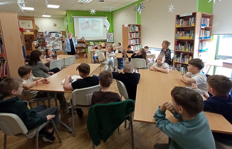 Kolorowe pomieszczenie, przy prostokątnych stołach stołach, na krzesłach siedzą dzieci przed dziećmi stoi kobieta prowadząca zajęcia.