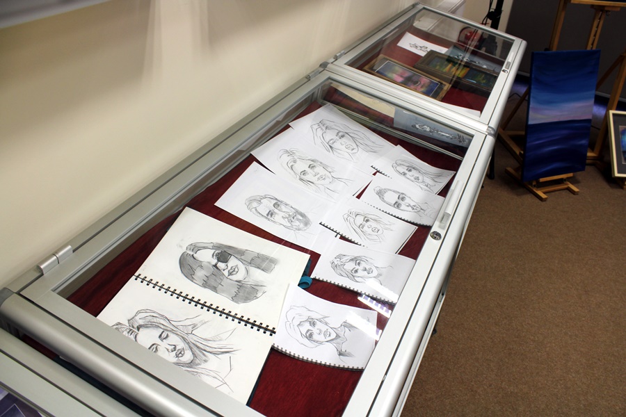 Prace artystki ułożone w gablotach, prace przedstawiają szkice twarzy