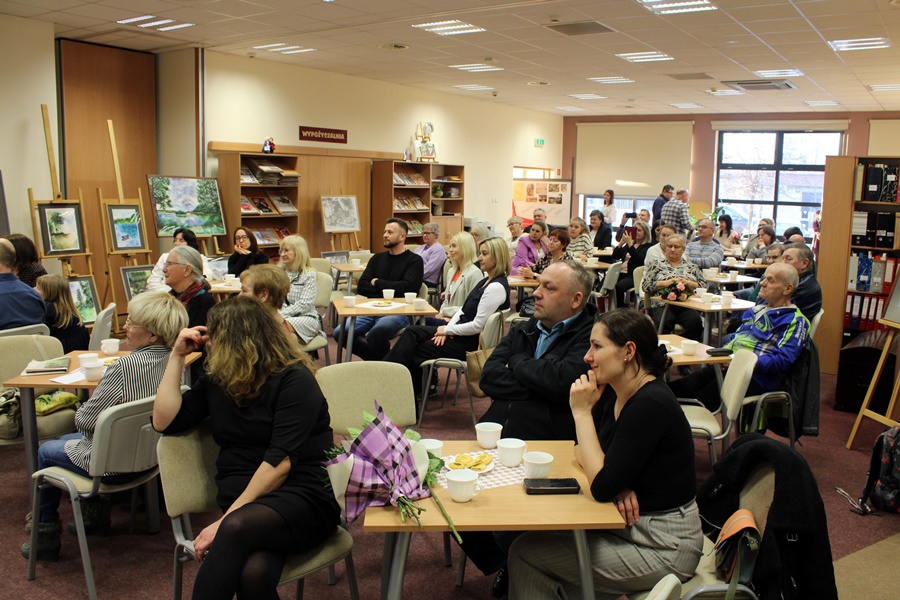 Goście obecni na spotkaniu z uwagą słuchają rozmowy prowadzonej z zaproszonym gościem, widzowie siedzą na krzesłach przy kwadratowych stolikach na których leżą talerzyki z ciastkami i stoją filiżanki z kawą lub herbatą