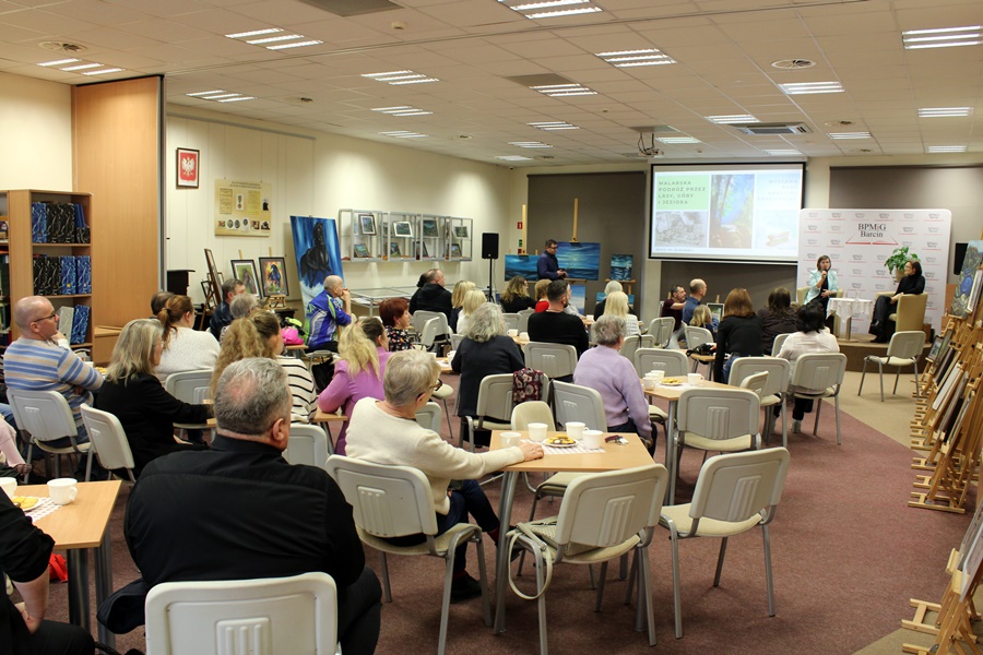 Goście obecni na spotkaniu z uwagą słuchają rozmowy prowadzonej z zaproszonym gościem, widzowie siedzą na krzesłach przy kwadratowych stolikach na których leżą talerzyki z ciastkami i stoją filiżanki z kawą lub herbatą
