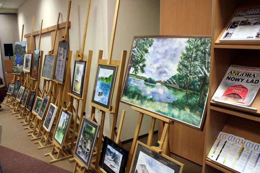 kolorowe obrazy ustawione wzdłuż ściany na sztalugach, wszytkie obrazy pzredstawiają różnego rodzaju krajobrazy