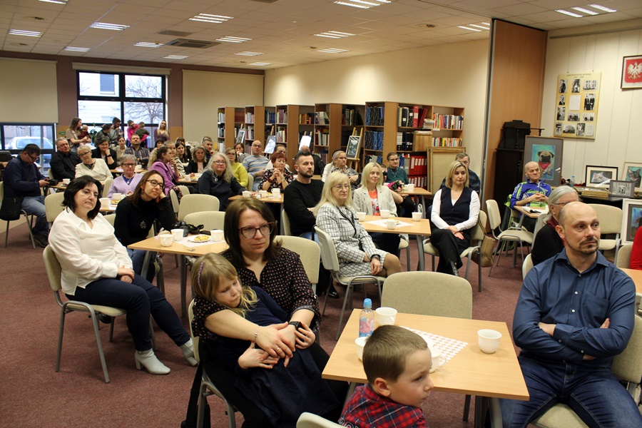 Goście obecni na spotkaniu z uwagą słuchają rozmowy prowadzonej z zaproszonym gościem, widzowie siedzą na krzesłach przy kwadratowych stolikach na których leżą talerzyki z ciastkami i stoją filiżanki z kawą lub herbatą