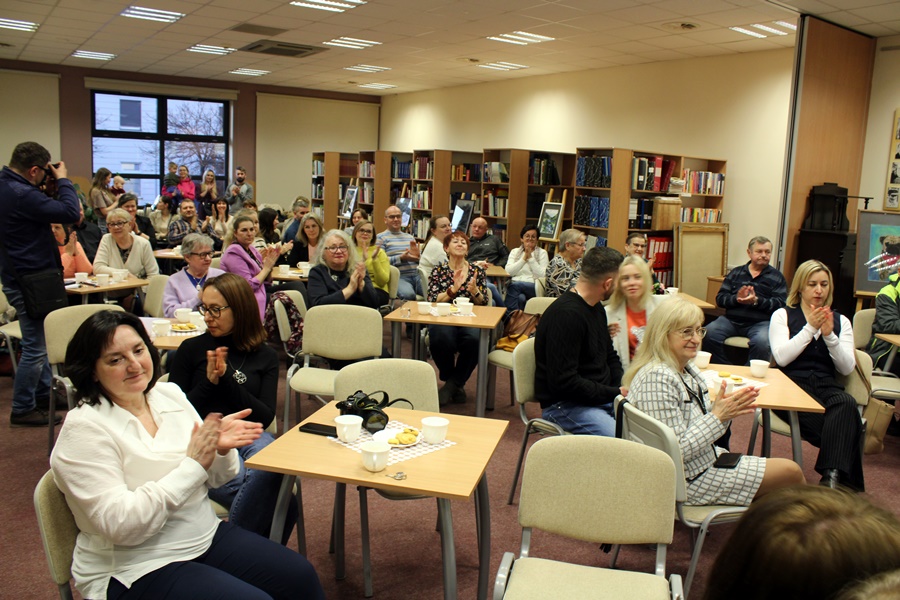 Goście obecni na spotkaniu z uwagą słuchają rozmowy prowadzonej z zaproszonym gościem, widzowie siedzą na krzesłach przy kwadratowych stolikach na których leżą talerzyki z ciastkami i stoją filiżanki z kawą lub herbatą