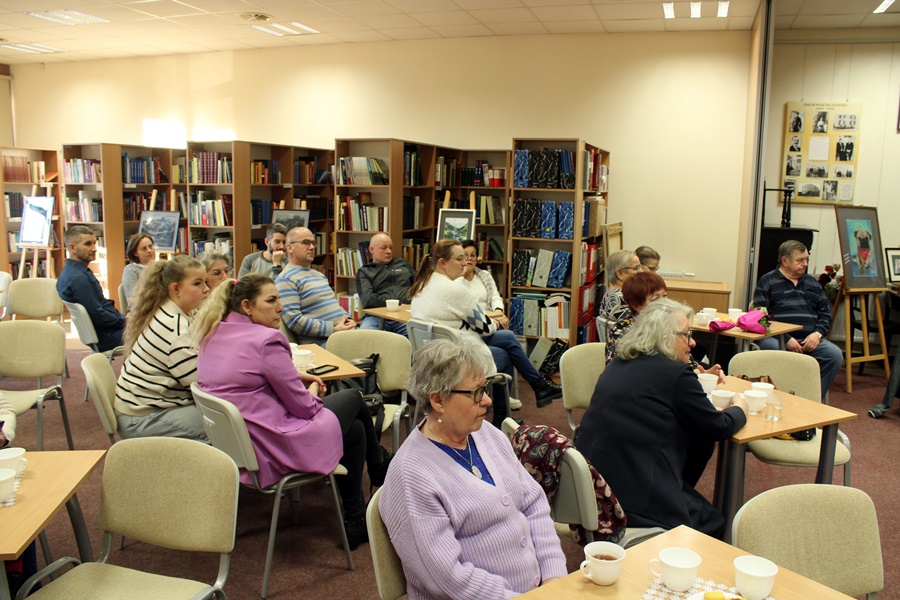 Goście obecni na spotkaniu z uwagą słuchają rozmowy prowadzonej z zaproszonym gościem, widzowie siedzą na krzesłach przy kwadratowych stolikach na których leżą talerzyki z ciastkami i stoją filiżanki z kawą lub herbatą