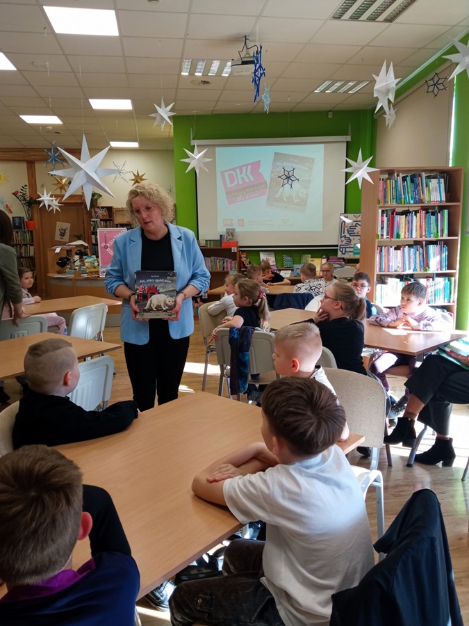 kobieta stoi pomiędzy stołami przy których siedzą dzieci, w rękach trzyma kolorową książkę