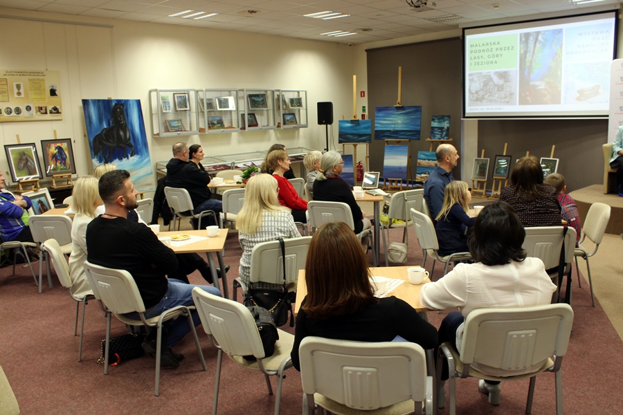 Goście obecni na spotkaniu z uwagą słuchają rozmowy prowadzonej z zaproszonym gościem, widzowie siedzą na krzesłach przy kwadratowych stolikach na których leżą talerzyki z ciastkami i stoją filiżanki z kawą lub herbatą