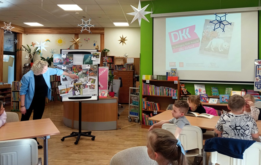 kobieta stoi przed grupą dzieci siedzących przy stołach i rozmawia z nimi