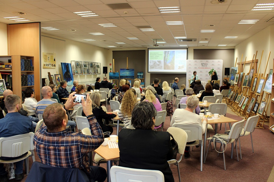 Goście obecni na spotkaniu z uwagą słuchają rozmowy prowadzonej z zaproszonym gościem, widzowie siedzą na krzesłach przy kwadratowych stolikach na których leżą talerzyki z ciastkami i stoją filiżanki z kawą lub herbatą