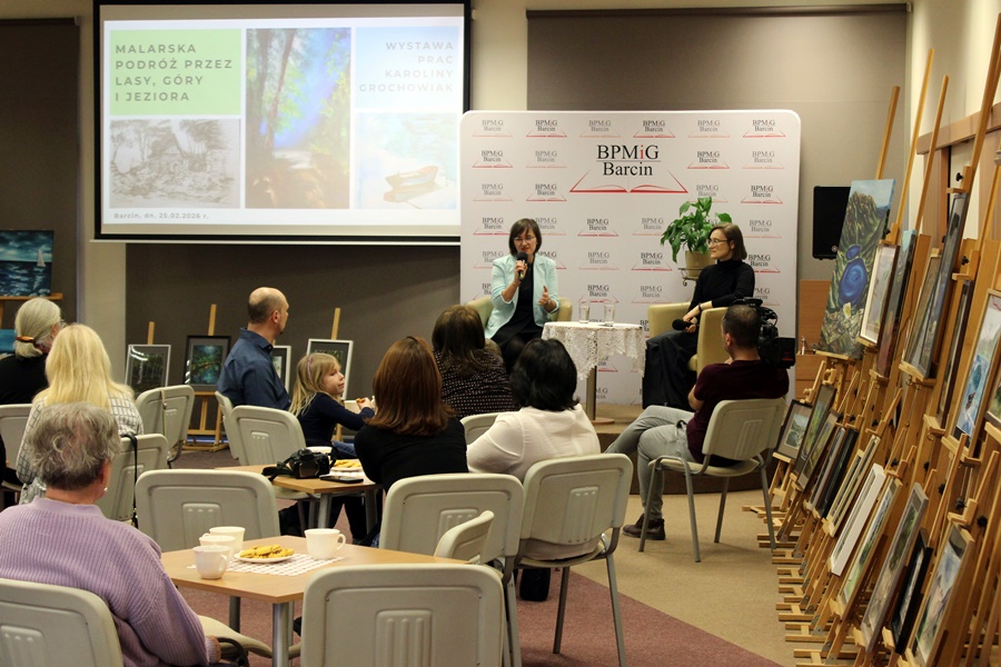 W głębi widać siedzące na scenie prowadzącą spotkanie i zaproszoną artystkę, bliżej widoczna jest cześć publiczności