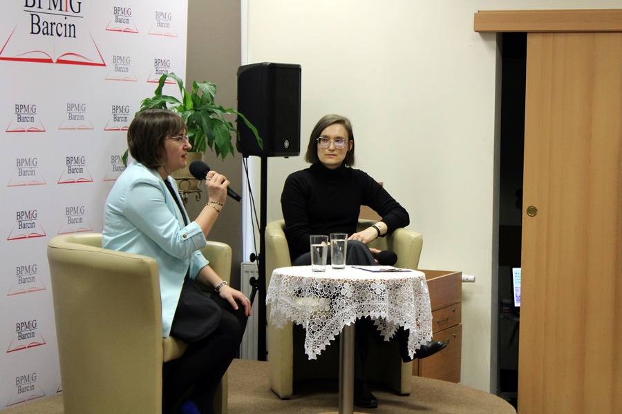 Prowadząca spotkanie bibliotekarka i zaproszona artystka siedzą w fotelach na scenie i rozmawiają między sobą