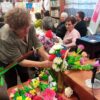 Kobieta tworzy kompozycję kwiatową w wazonie inne osoby przyglądają się jej pracy
