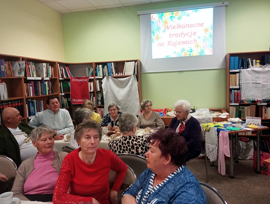 Uczestnicy spotkania siedzą wokół prostokątnych stołów