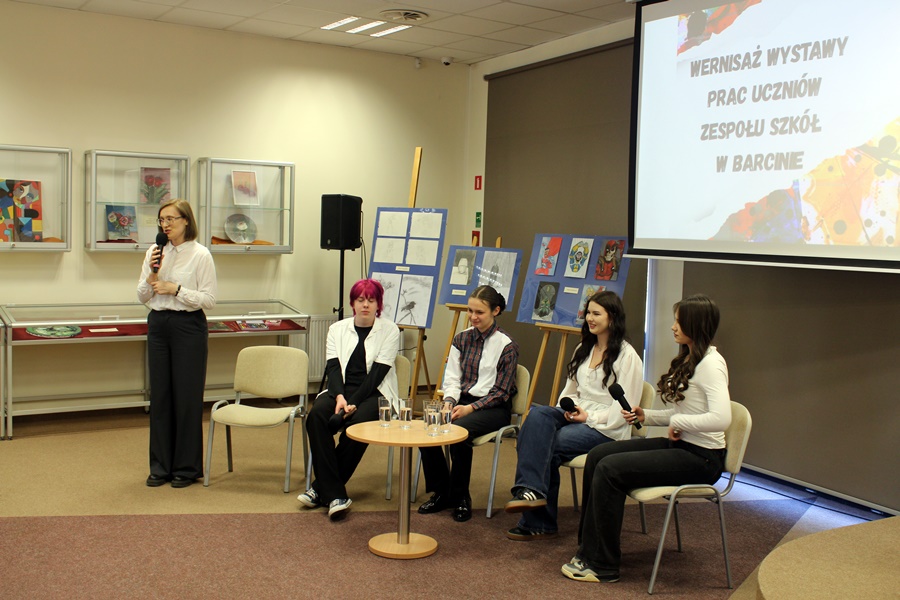 Prowadząca spotkanie stoi i wita zebraną publiczność a obok niej siedzą artyści których dotyczywystawapra