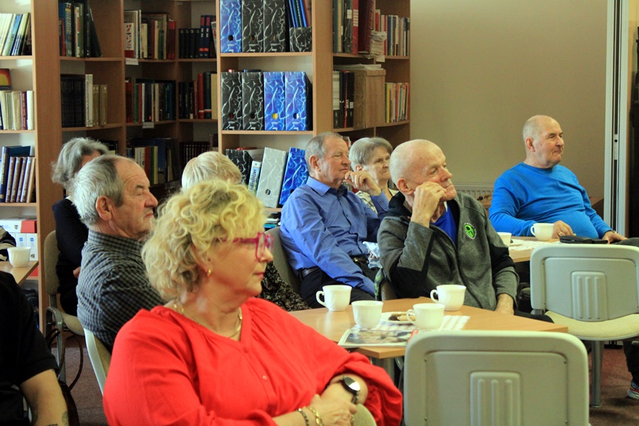 ludzie siedzą na krzesłach przy kwadratowych stolikach i słuchają rozmowy prowadzonej z zaproszoną gościnią.