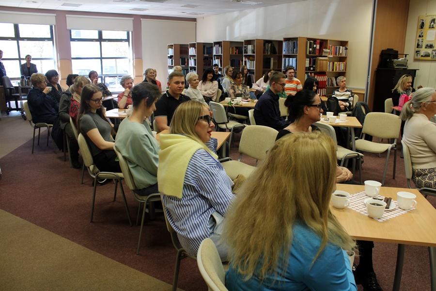 Zdjęcie publiczności, zgromadzeni goście siedzą na krzesłach przy kwadratowych stolikach, popijając kawę lub herbatę