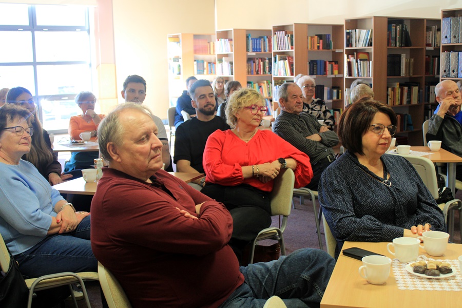 ludzie siedzą na krzesłach przy kwadratowych stolikach i słuchają rozmowy prowadzonej z zaproszoną gościnią.
