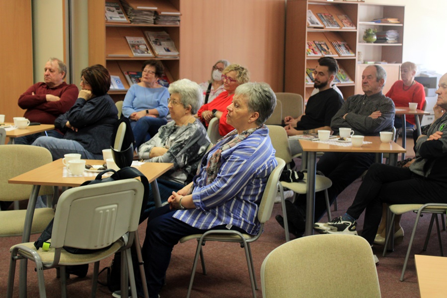 ludzie siedzą na krzesłach przy kwadratowych stolikach i słuchają rozmowy prowadzonej z zaproszoną gościnią.