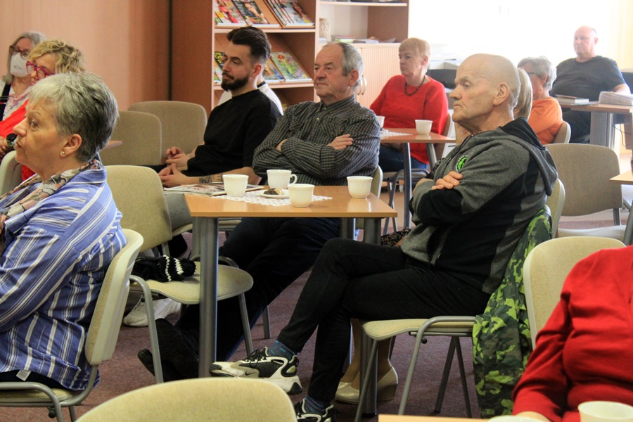 ludzie siedzą na krzesłach przy kwadratowych stolikach i słuchają rozmowy prowadzonej z zaproszoną gościnią.
