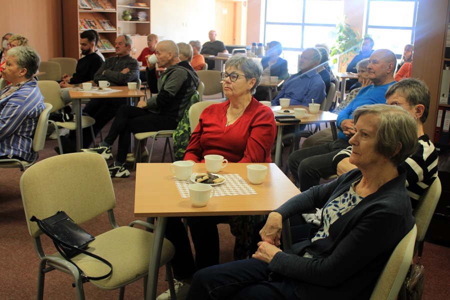 ludzie siedzą na krzesłach przy kwadratowych stolikach i słuchają rozmowy prowadzonej z zaproszoną gościnią.