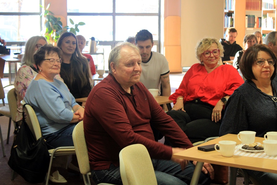 ludzie siedzą na krzesłach przy kwadratowych stolikach i słuchają rozmowy prowadzonej z zaproszoną gościnią.