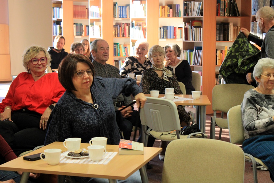 ludzie siedzą na krzesłach przy kwadratowych stolikach i słuchają rozmowy prowadzonej z zaproszoną gościnią.