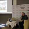 zaproszony autor siedzi na scenie, mówi do mikrofonu i przełącza przygotowaną prezentację