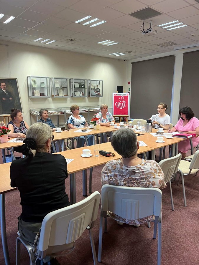 Uczestniczki spotkania i zaproszona bibliotekarka siedzą przy stołach i rozmawiają o ksiażkach.