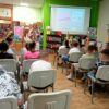 Dzieci siedzą na krzesłach a bibliotekarka chodzi pomiędzy nimi.