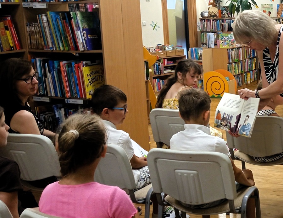Bibliotekarka pokazuje jednemu z dzieci zawartość książki o której opowiada.